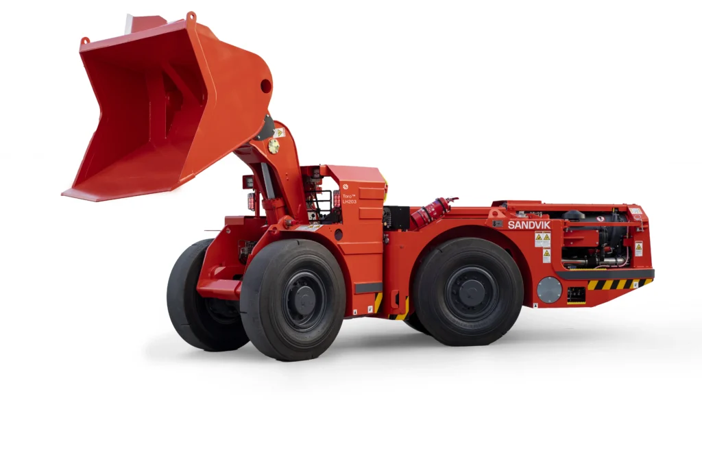 Sandvik underground mining loader shown in an industrial mine setting from a front-side angle. The heavy-duty yellow LHD machine features a large bucket, articulated frame, and rugged tires designed for loading, hauling, and transporting material in underground mining operations.