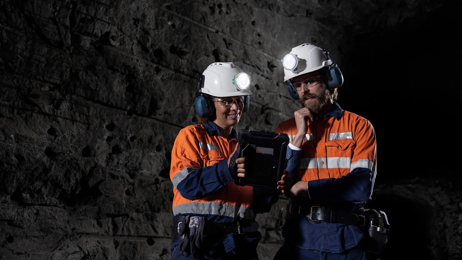 Group of miners in a tunnel inside an underground mine, wearing high-visibility protective clothing and helmets with headlamps. They are standing beside a Sandvik mining machine, observing operations in a dimly lit, industrial underground environment.